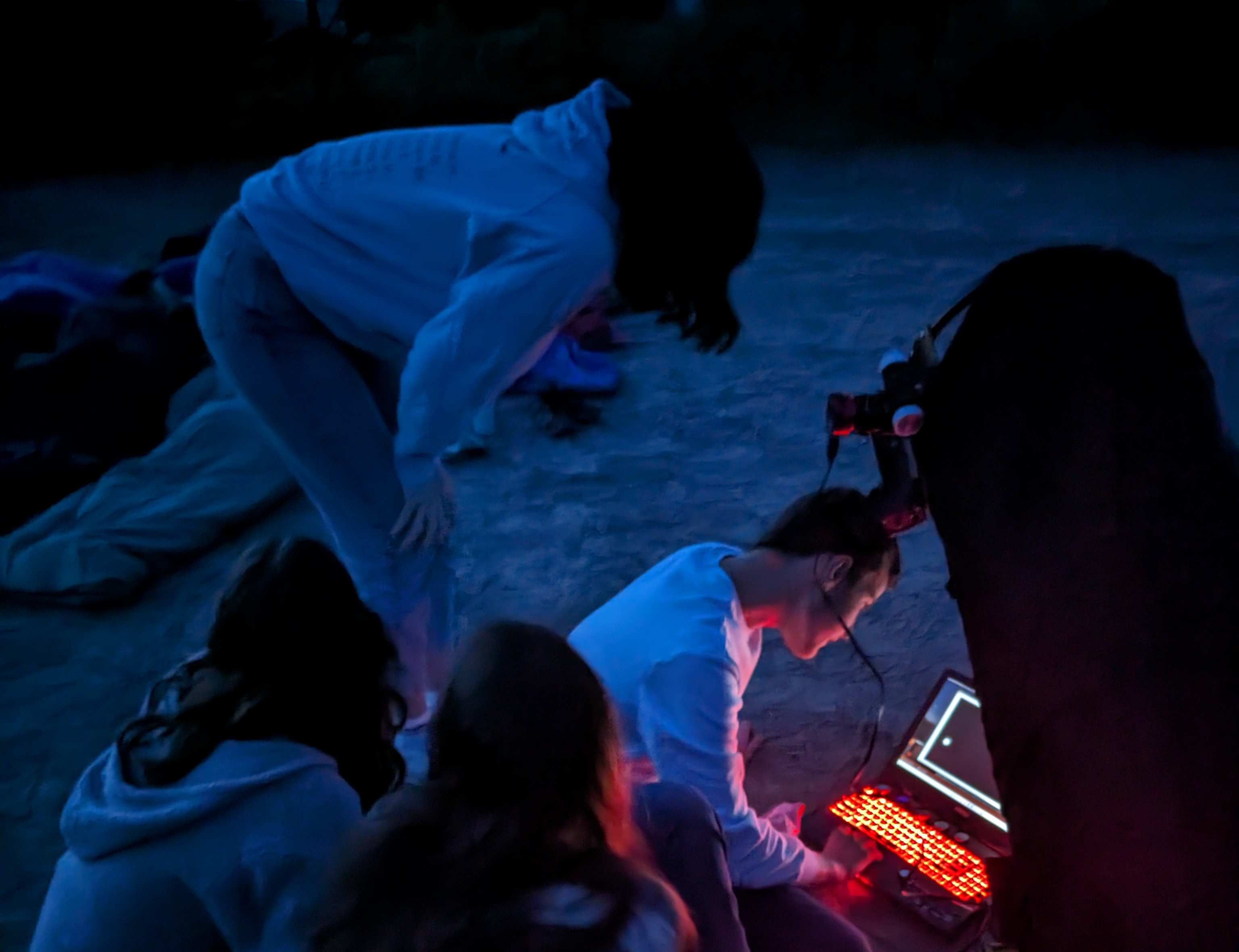 A participant hunched over a leader at a laptop on the dirt ground in dim evening light.